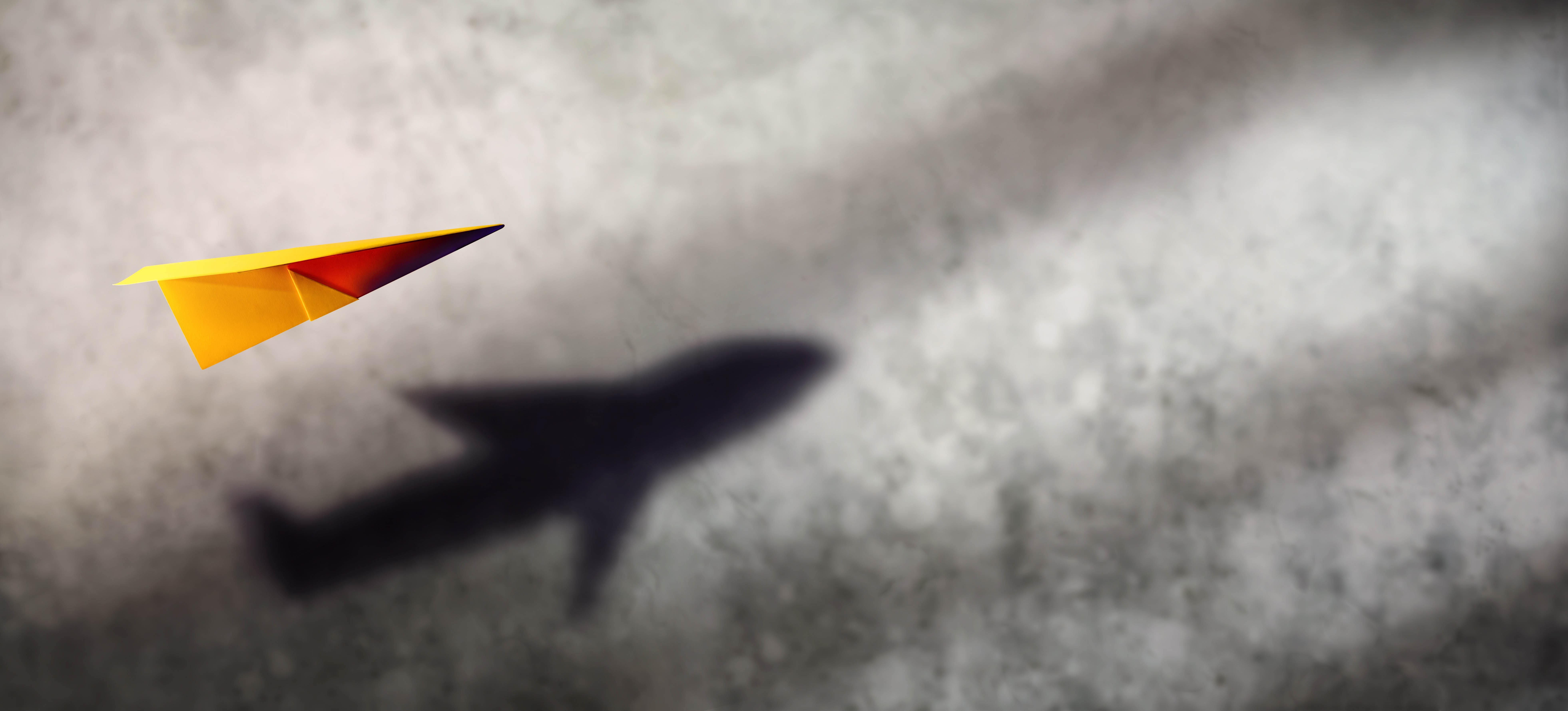 Yellow paper plane flies over a gray background, the shadow is of an actual airplane Yellow paper plane flies over a gray background, the shadow is of an actual airplane